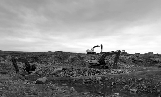 Excavators working a stripped, rock-strewn site under an overcast sky, standing water in the foreground