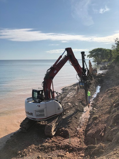 Pilon excavator and drill rig working shoreline stabilization at the water's edge